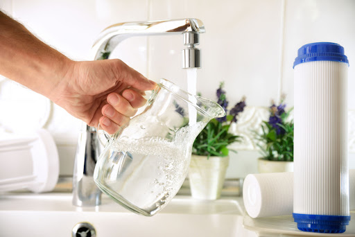 A person filling a pitcher of water from a kitchen sink faucet with a water filter situation nearby.