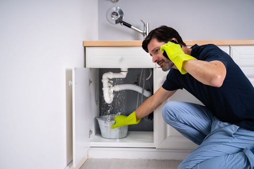 A man wearing gloves holding a bucket under a leaky sink pipe while calling someone on the phone.